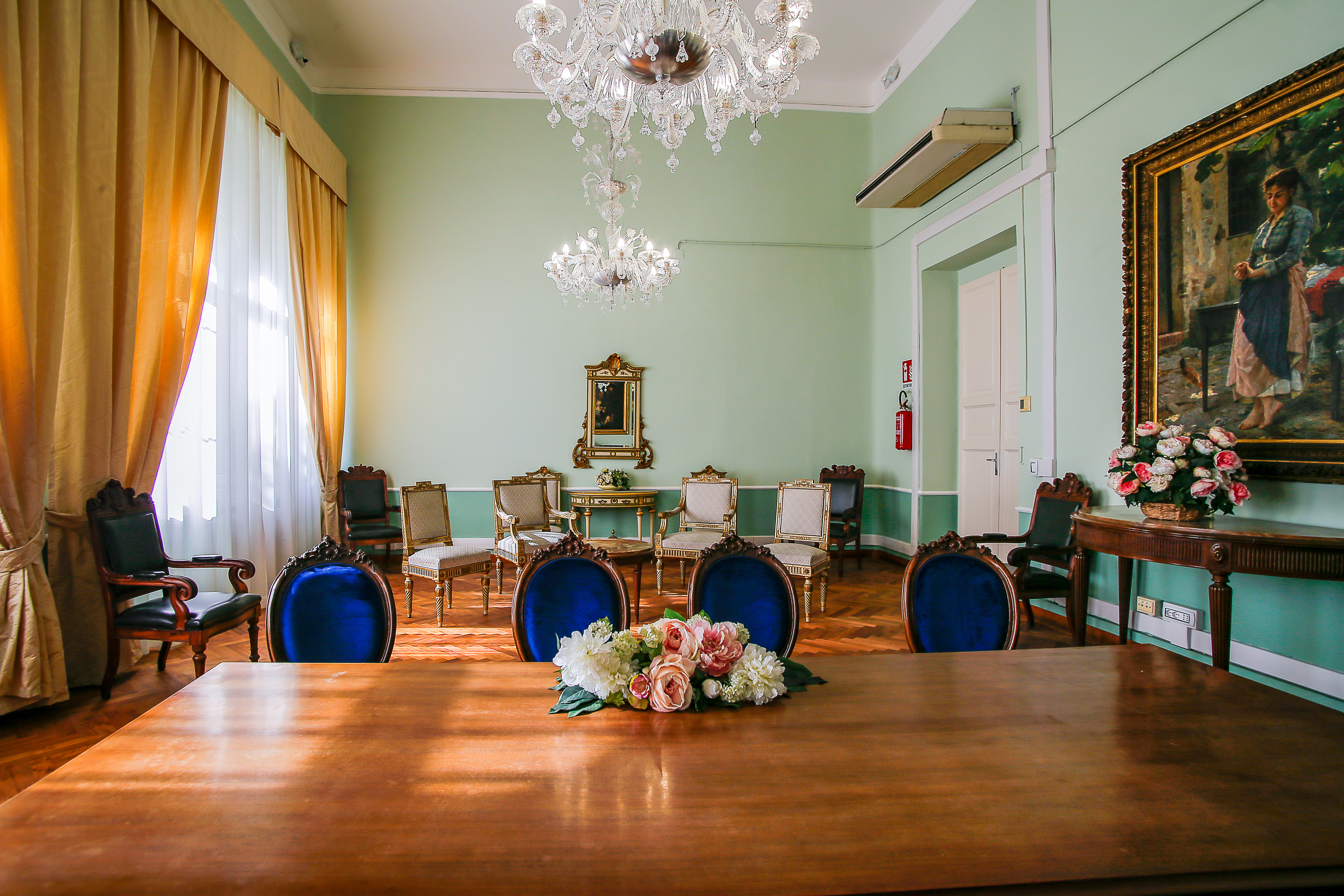 Sala cerimoniale elegante con arredi d’epoca, pareti verde pastello, pavimento in parquet e ampie tende dorate su una finestra luminosa. Al centro della scena, un tavolo in legno con composizione floreale, sedie imbottite con velluto blu, lampadari in cristallo e quadri d’arte classica alle pareti.