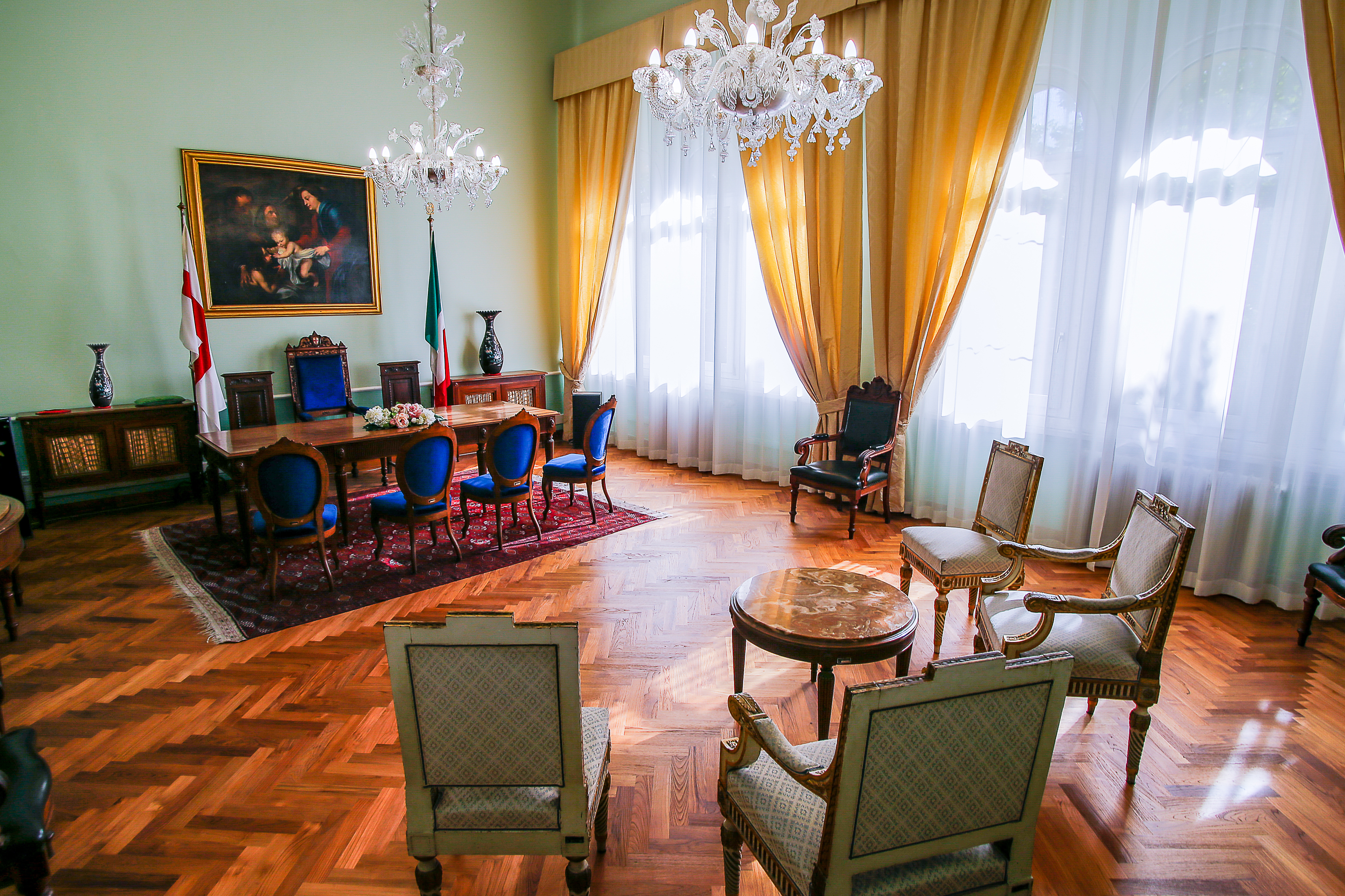 Sala cerimonie presso la sede di Corso Torino, con arredi eleganti in stile classico, sedie imbottite blu e beige, grandi finestre con tende dorate, tavolo centrale in legno con addobbo floreale, bandiere istituzionali e lampadari in cristallo.