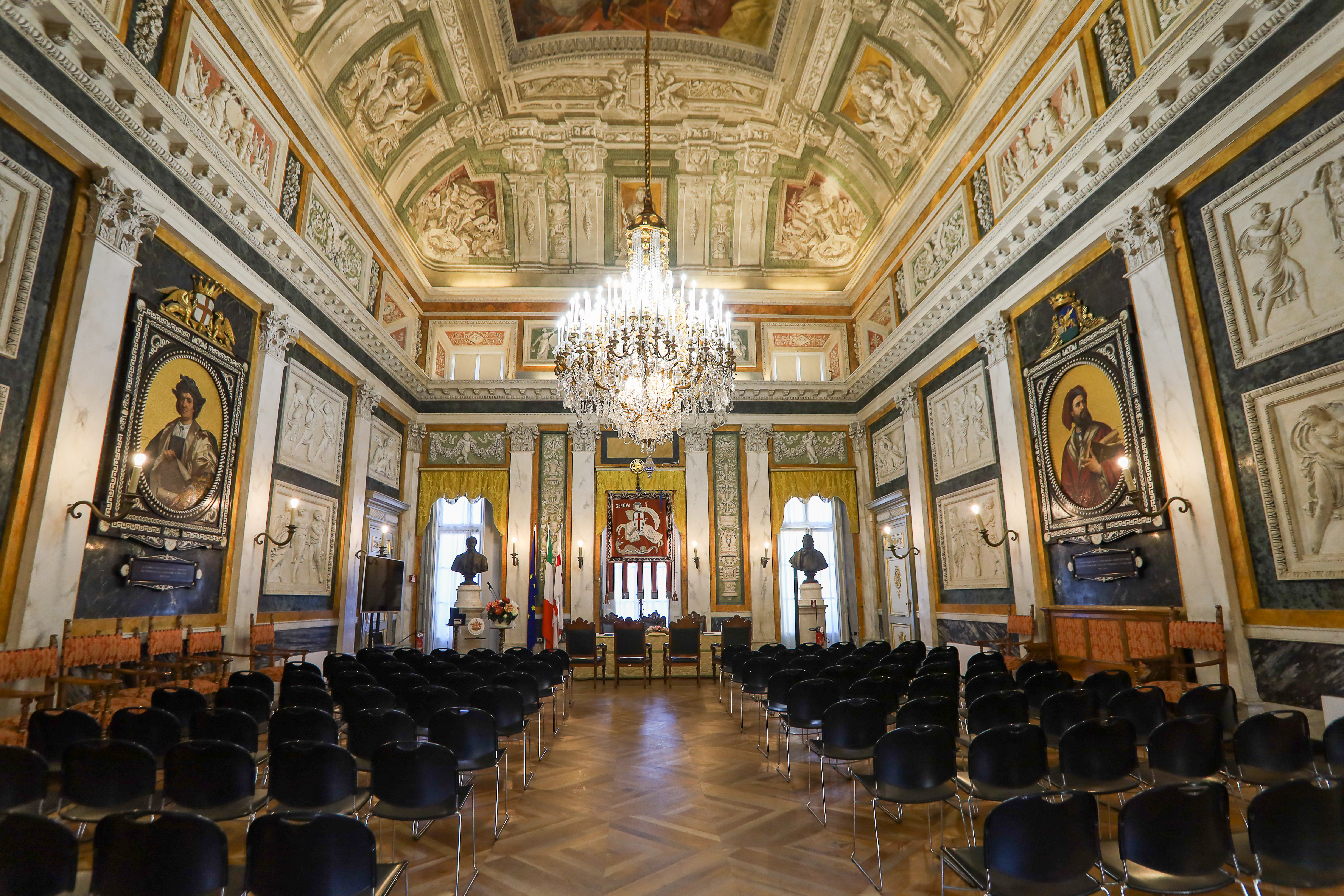 Salone di rappresentanza del Comune di Genova, con tavolo cerimoniale, sedie in legno, decorazioni storiche e bandiere istituzionali.
