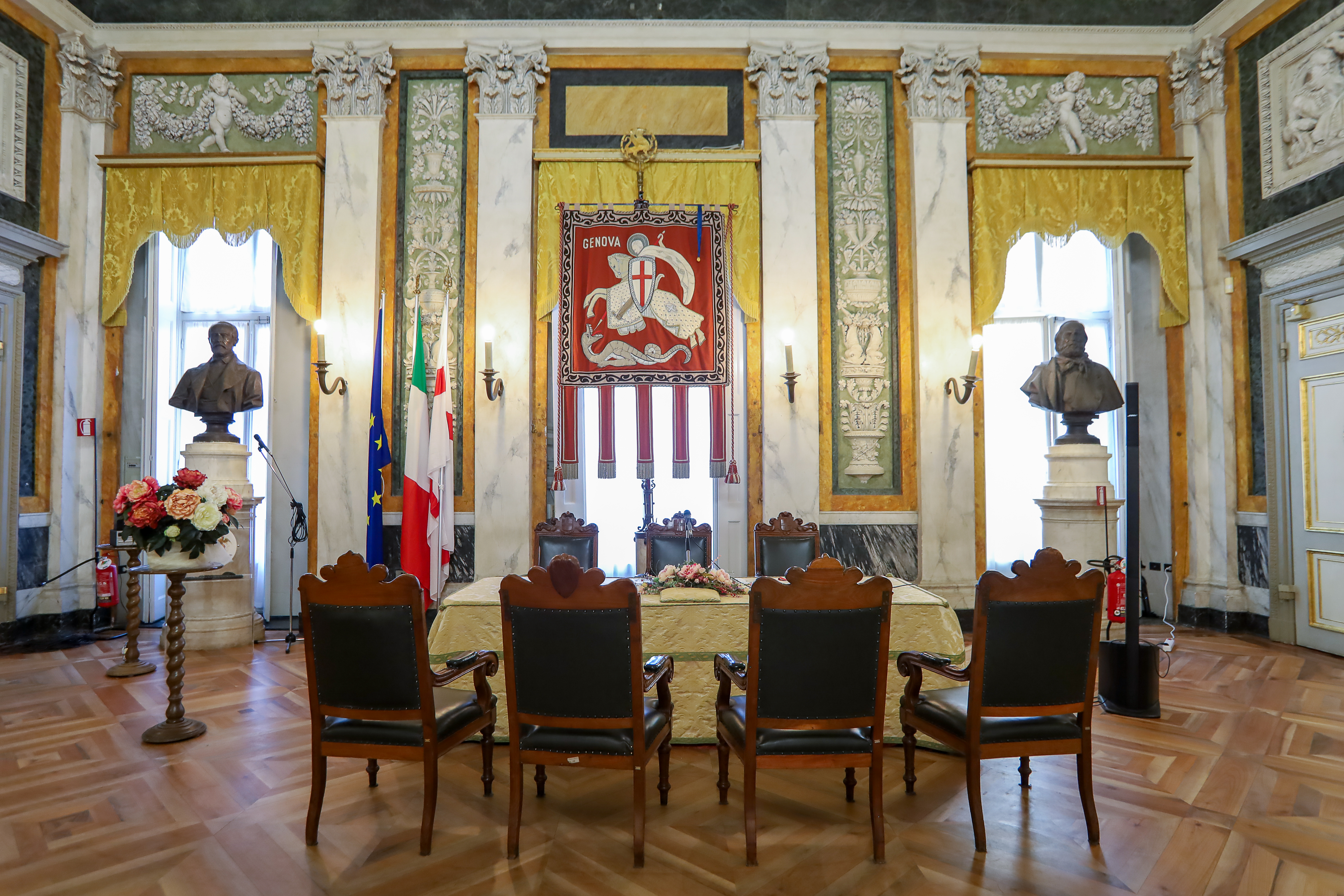 Salone di rappresentanza del Comune di Genova, ambiente solenne con pareti decorate in stile neoclassico, colonne marmoree, busti commemorativi, bandiere istituzionali e il gonfalone della città al centro. Tavolo cerimoniale rivestito da tessuto damascato, con sedute in legno scuro e dettagli dorati. Il pavimento in parquet e i tendaggi color oro completano l’atmosfera storica.
