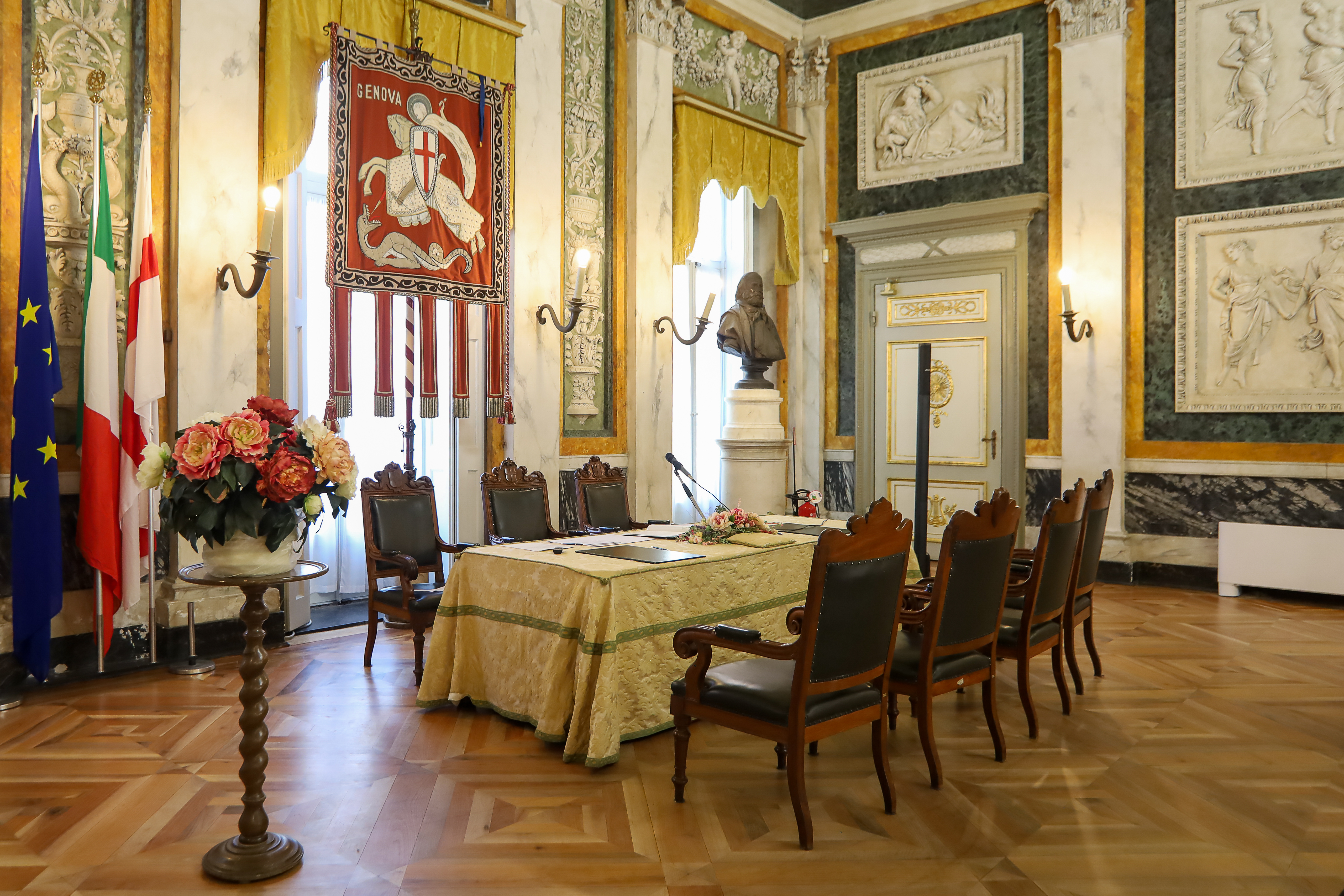 Salone di rappresentanza del Comune di Genova, ambiente solenne con pareti decorate in stile neoclassico, colonne marmoree, busti commemorativi, bandiere istituzionali e il gonfalone della città al centro. Tavolo cerimoniale rivestito da tessuto damascato, con sedute in legno scuro e dettagli dorati. Il pavimento in parquet e i tendaggi color oro completano l’atmosfera storica.
