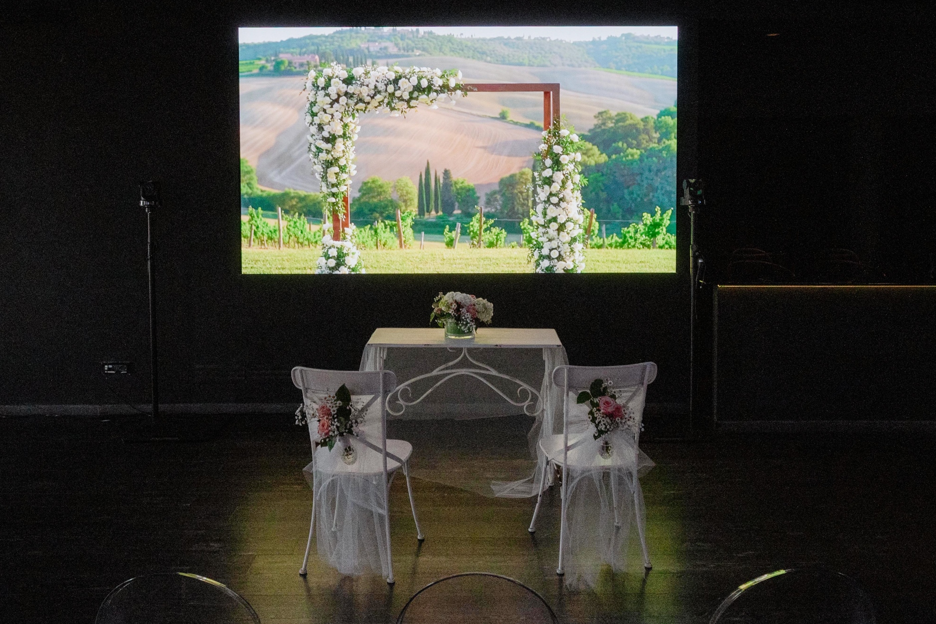 Allestimento per cerimonia matrimoniale nella Terrazza Colombo: due sedie decorate con tulle e fiori bianchi davanti a un tavolo nuziale, con sullo sfondo una grande proiezione di paesaggio naturale
