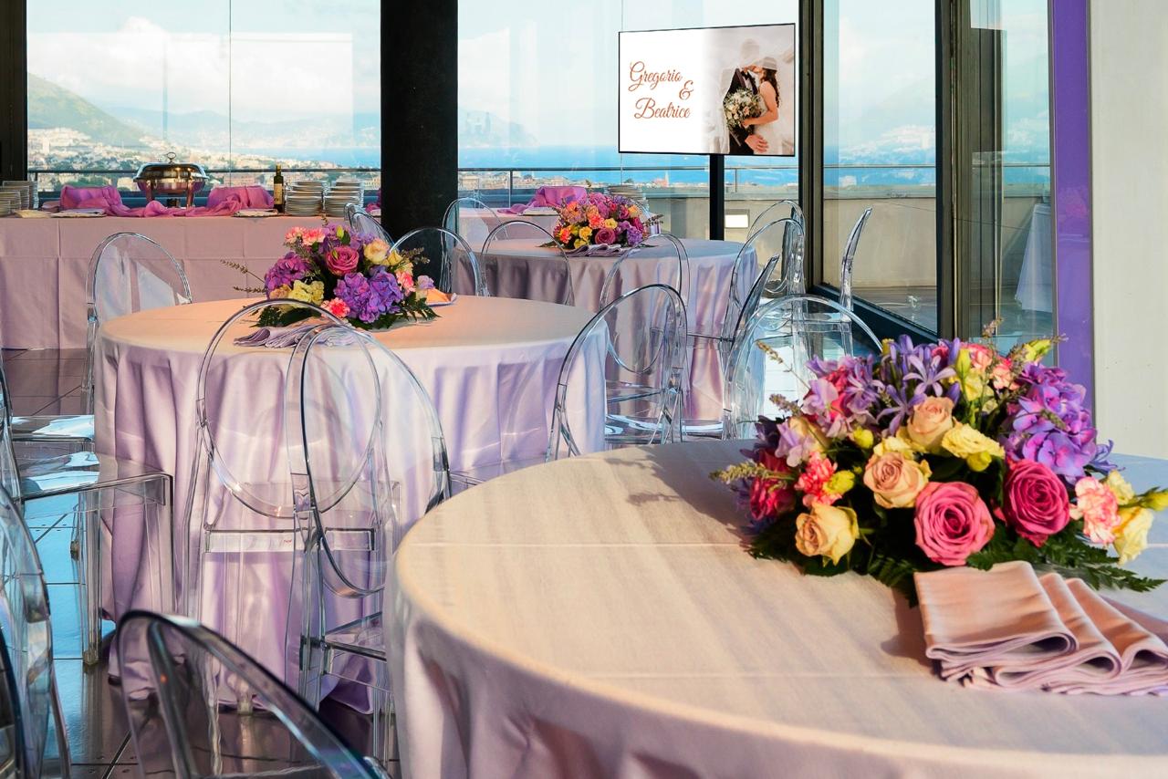 Sala allestita per ricevimento nella Terrazza Colombo: tavoli rotondi con tovaglie color lavanda, sedie trasparenti e centrotavola floreali con vista panoramica su Genova e il mare