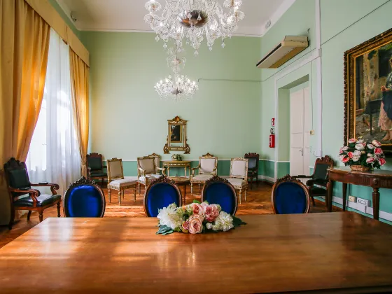 Sala cerimoniale elegante con arredi d’epoca, pareti verde pastello, pavimento in parquet e ampie tende dorate su una finestra luminosa. Al centro della scena, un tavolo in legno con composizione floreale, sedie imbottite con velluto blu, lampadari in cristallo e quadri d’arte classica alle pareti.