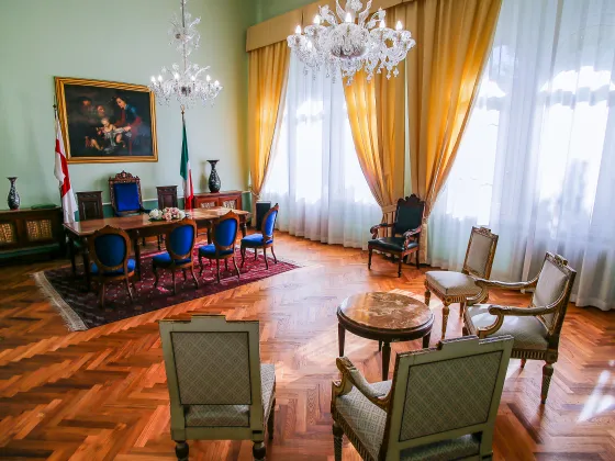 Sala cerimonie presso la sede di Corso Torino, con arredi eleganti in stile classico, sedie imbottite blu e beige, grandi finestre con tende dorate, tavolo centrale in legno con addobbo floreale, bandiere istituzionali e lampadari in cristallo.