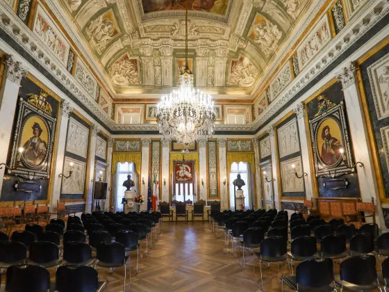 Salone di rappresentanza del Comune di Genova, con tavolo cerimoniale, sedie in legno, decorazioni storiche e bandiere istituzionali.