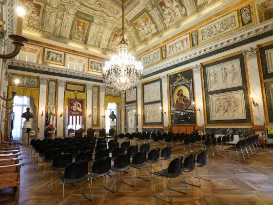 Salone di rappresentanza del Comune di Genova, con tavolo cerimoniale, sedie in legno, decorazioni storiche e bandiere istituzionali.