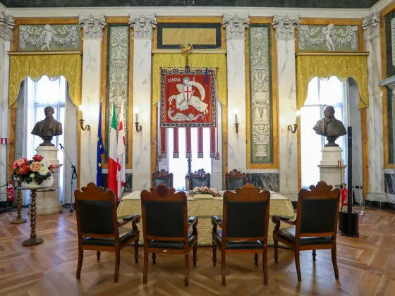 Salone di rappresentanza del Comune di Genova, ambiente solenne con pareti decorate in stile neoclassico, colonne marmoree, busti commemorativi, bandiere istituzionali e il gonfalone della città al centro. Tavolo cerimoniale rivestito da tessuto damascato, con sedute in legno scuro e dettagli dorati. Il pavimento in parquet e i tendaggi color oro completano l’atmosfera storica.