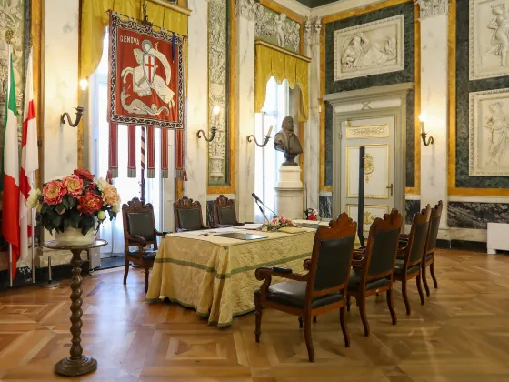 Salone di rappresentanza del Comune di Genova, ambiente solenne con pareti decorate in stile neoclassico, colonne marmoree, busti commemorativi, bandiere istituzionali e il gonfalone della città al centro. Tavolo cerimoniale rivestito da tessuto damascato, con sedute in legno scuro e dettagli dorati. Il pavimento in parquet e i tendaggi color oro completano l’atmosfera storica.